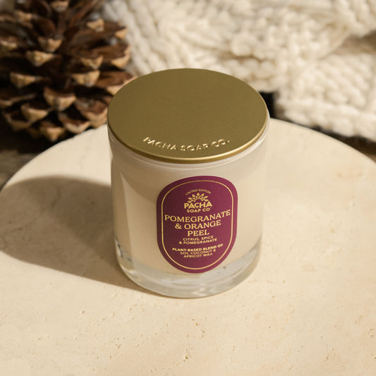 Pacha Soap's Pomegranate & Orange Peel Signature holiday candle, with a gold lid and a red label with gold foil and on a beautiful circle marble pedestal, next to a pinecone and knitted blanket.