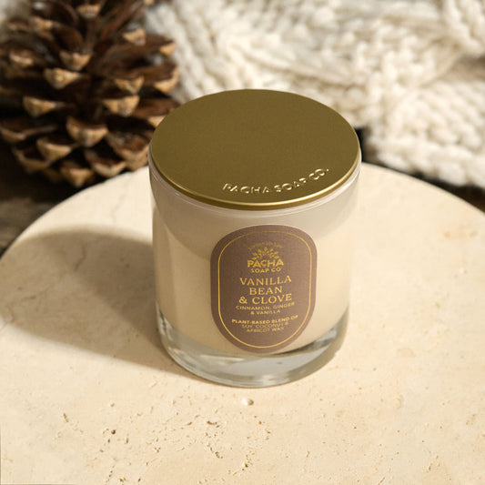 Pacha Soap's Vanilla Bean & Clove Signature holiday candle, with a gold lid and a red label with gold foil and on a beautiful circle marble pedestal, next to a pinecone and knitted blanket.