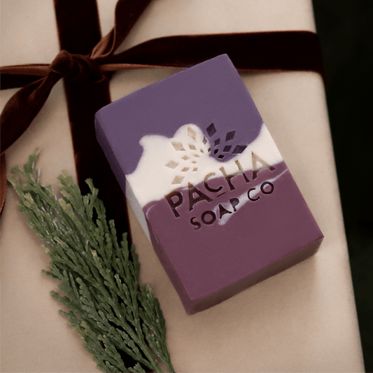 Pacha Soap Co.'s Pomegranate & Orange Peel purple, magenta, and cream bar soap on kraft paper and next to a brown velvet ribbon and pine sprig.