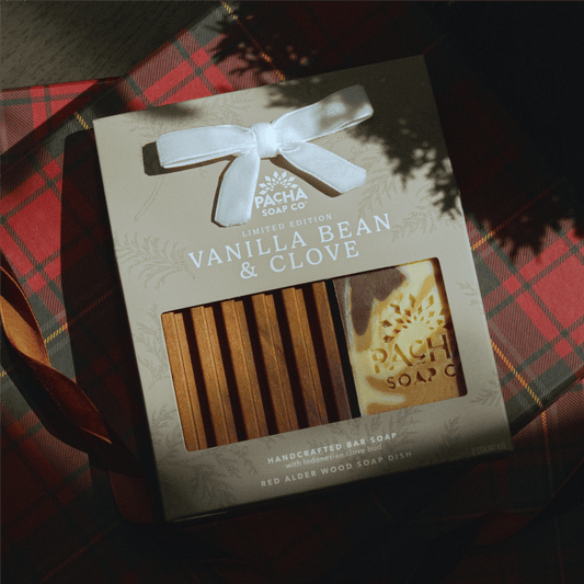 Vanilla Bean & Clove Dish set in a creamy beige color sits on a plaid present and next to ribbon. The contents inside are a bar of soap and a wooden soap dish, the bar being handcrafted by Pacha Soap Co.