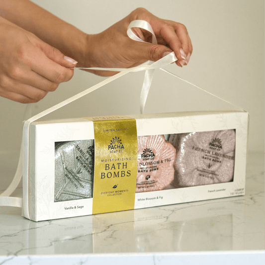 Two hands tying a white ribbon over Pacha Soap Co's Signature Bath bomb set with Vanilla & Sage, White Blossom & Fig, and French Lavender Bath bombs