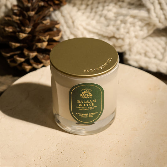 Pacha Soap's Balsam & Pine Signature holiday candle, with a gold lid and a red label with gold foil and on a beautiful circle marble pedestal, next to a pinecone and knitted blanket.