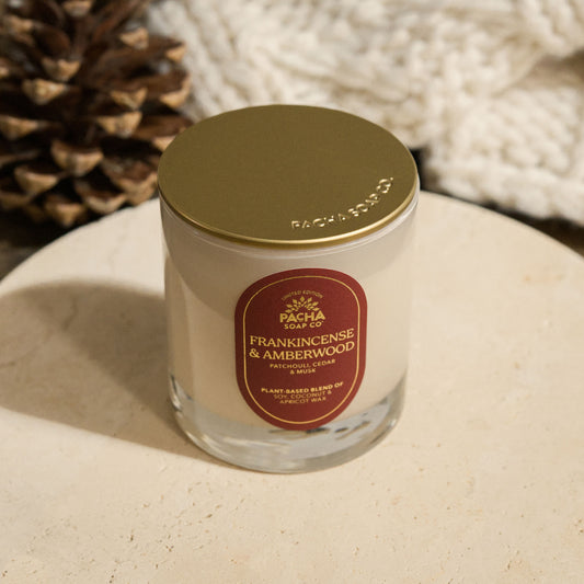 Pacha Soap's Frankincense & Amberwood Signature holiday candle, with a gold lid and a red label with gold foil and on a beautiful circle marble pedestal, next to a pinecone and knitted blanket.