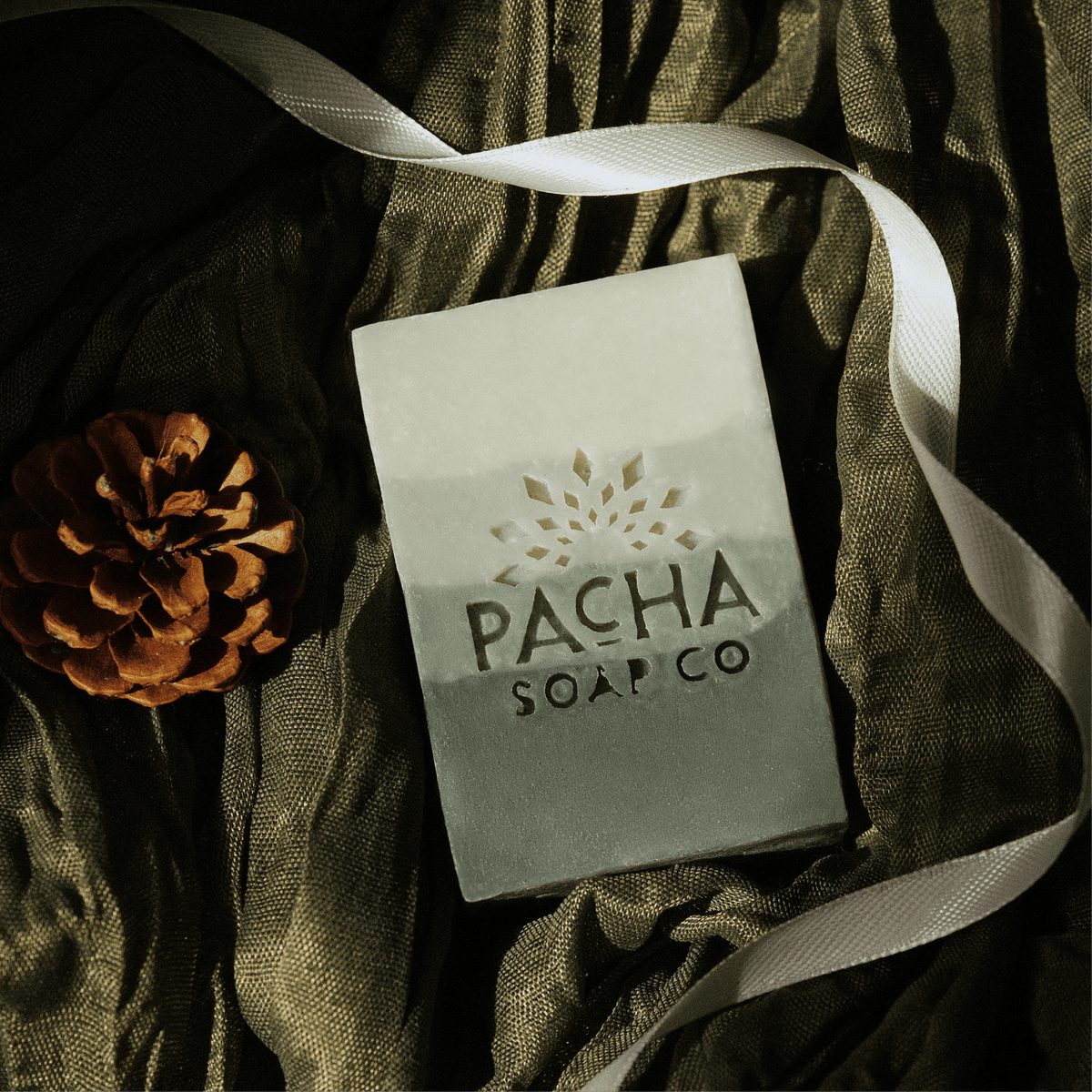 A rectangular bar of soap from Pacha Soap Co. lies on draped dark olive-green fabric, with a white ribbon and a brown pinecone nearby.