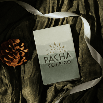 A rectangular bar of soap from Pacha Soap Co. lies on draped dark olive-green fabric, with a white ribbon and a brown pinecone nearby.