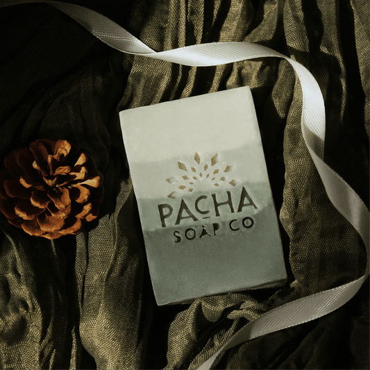 A rectangular bar of soap from Pacha Soap Co. lies on draped dark olive-green fabric, with a white ribbon and a brown pinecone nearby.