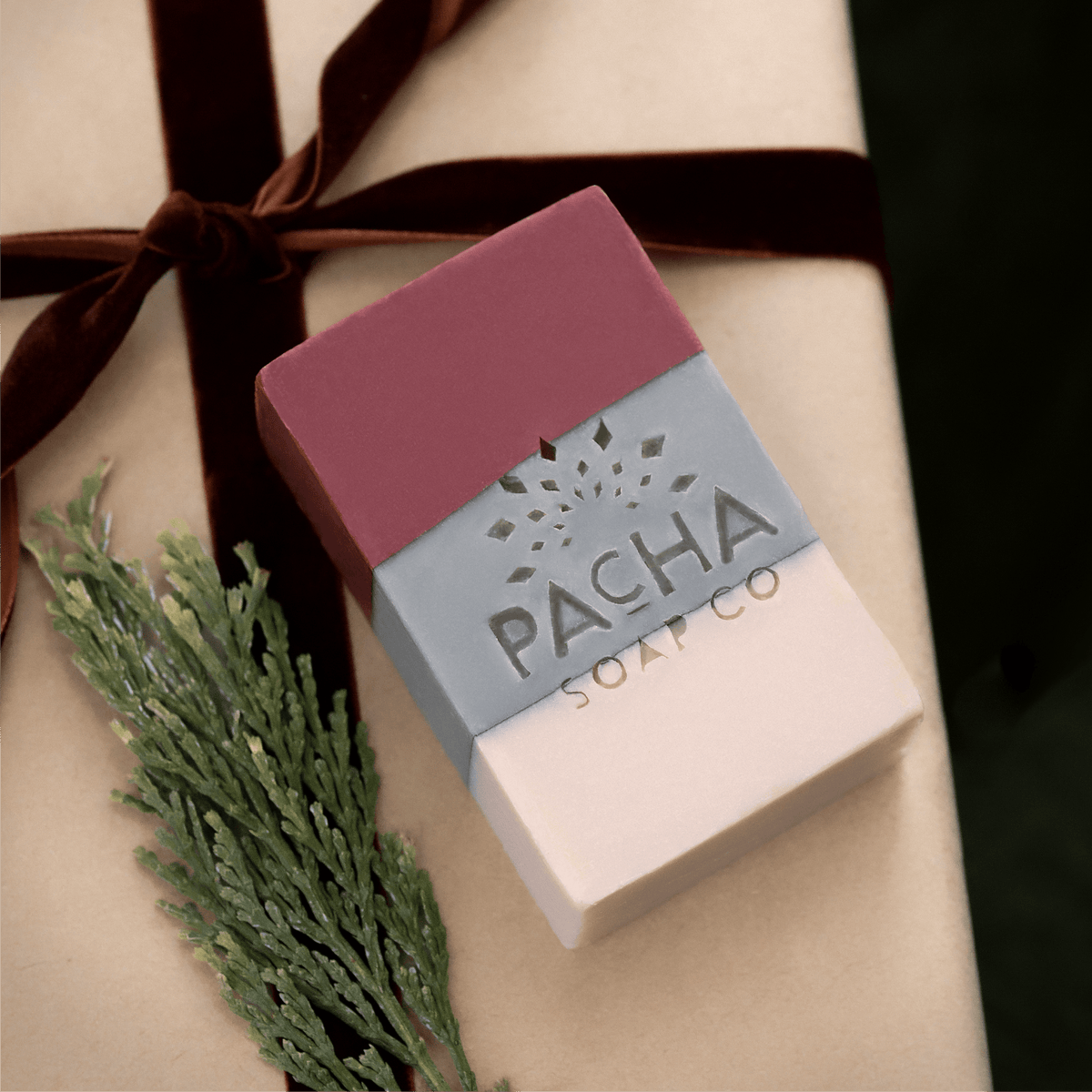 A rectangular bar of soap from Pacha Soap Co.  presented on a beige gift box tied with a dark brown ribbon and decorated with a green sprig.