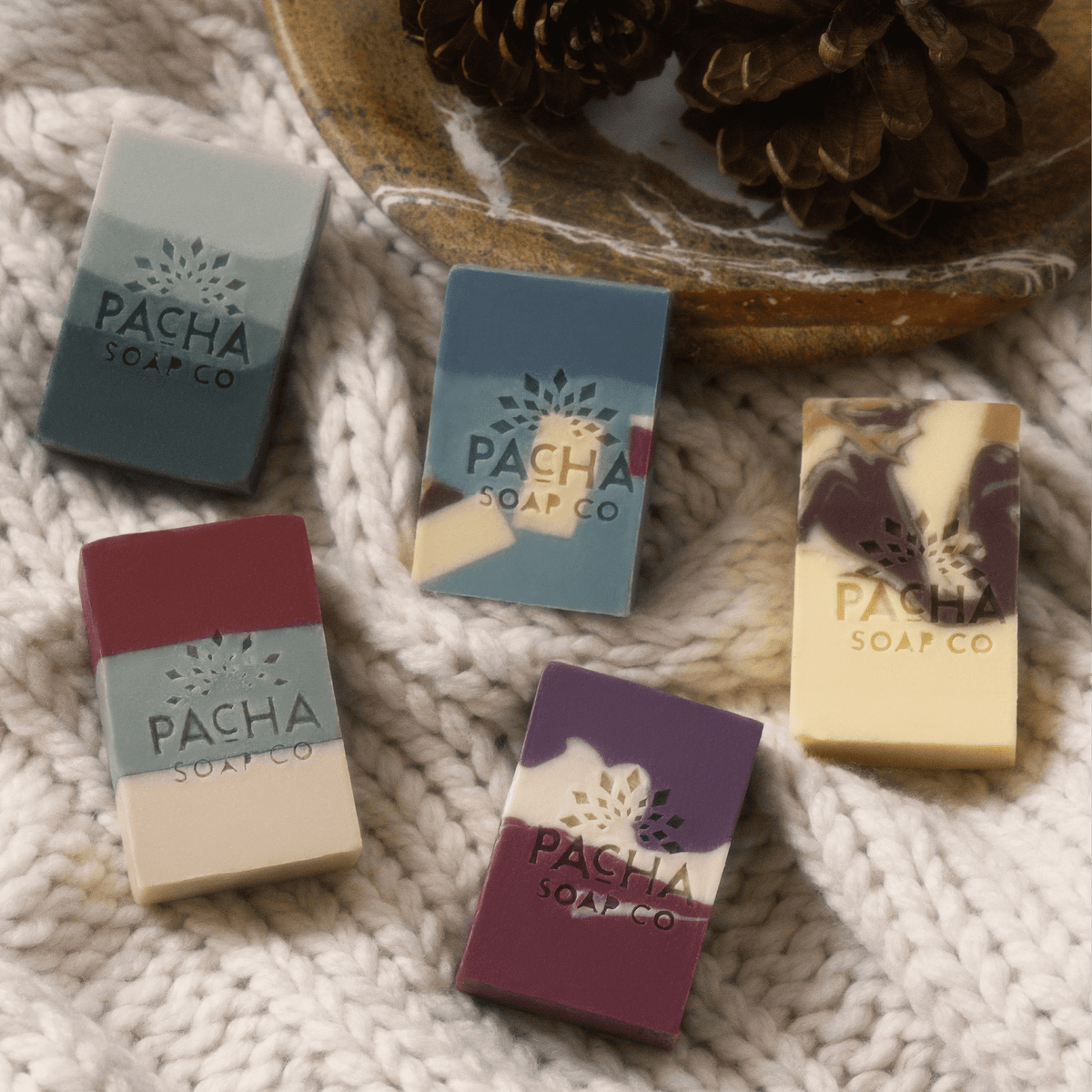 Five bars of Pacha Soap Co. artisan soap arranged on a knitted blanket next to a dish full of pinecones