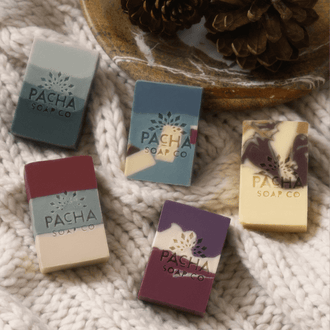 Five bars of Pacha Soap Co. artisan soap arranged on a knitted blanket next to a dish full of pinecones
