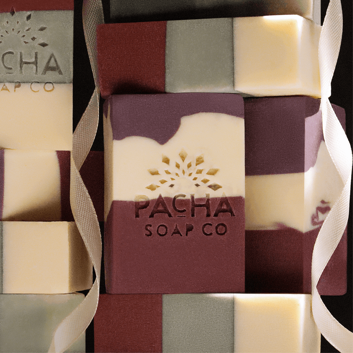 Abstract stack of Pacha Soap Co's Pomegranate & orange Peel and Frankincense & Amberwood bar soaps with a white ribbon draped over them.