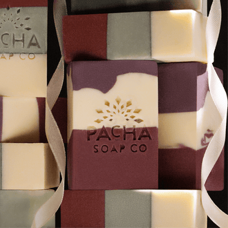 Abstract stack of Pacha Soap Co's Pomegranate & orange Peel and Frankincense & Amberwood bar soaps with a white ribbon draped over them.