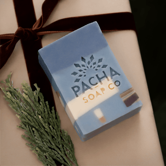 A rectangular bar of Pacha Soap Co. soap in a light blue and white marbled design, wrapped in a dark brown ribbon and presented on a beige gift box with a green pine sprig.