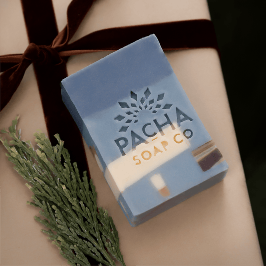 A rectangular bar of Pacha Soap Co. soap in a light blue and white marbled design, wrapped in a dark brown ribbon and presented on a beige gift box with a green pine sprig.