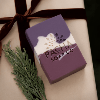 Pacha Soap Co.'s Pomegranate & Orange Peel purple, magenta, and cream bar soap on kraft paper and next to a brown velvet ribbon and pine sprig.