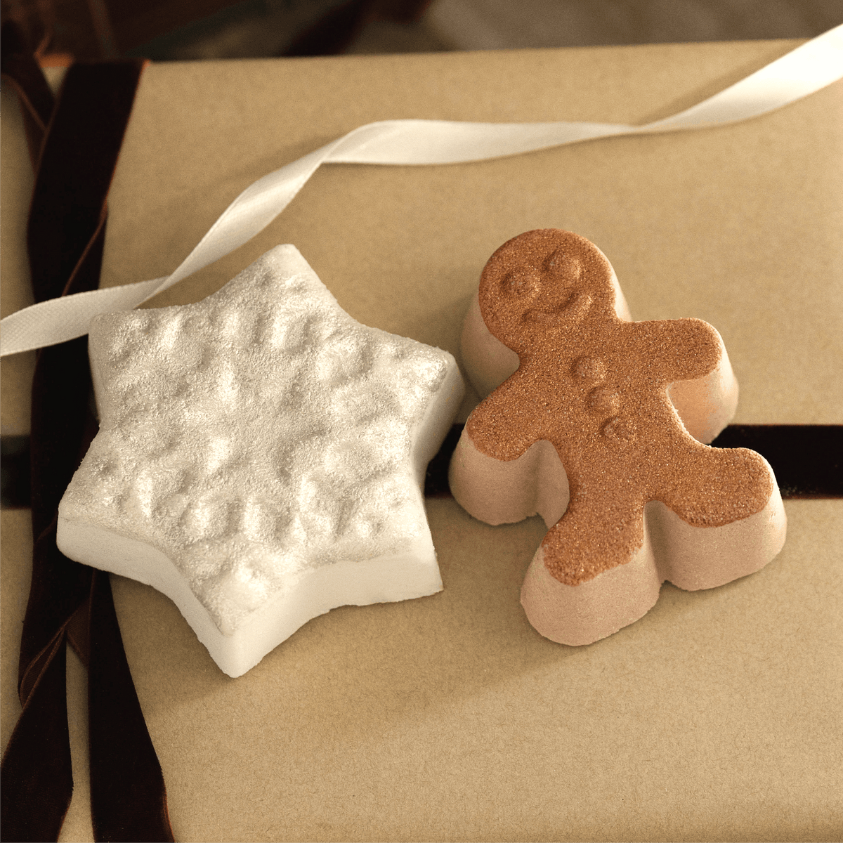 Two decorative bath bombs shaped like a white star and a gingerbread man, displayed on a brown kraft paper surface with a white ribbon.
