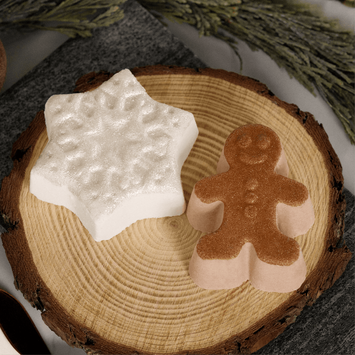 Two decorative bath bombs shaped like a white star and a gingerbread man, displayed on a round wood slice with dried botanicals in the background.