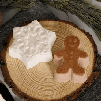 Two decorative bath bombs shaped like a white star and a gingerbread man, displayed on a round wood slice with dried botanicals in the background.