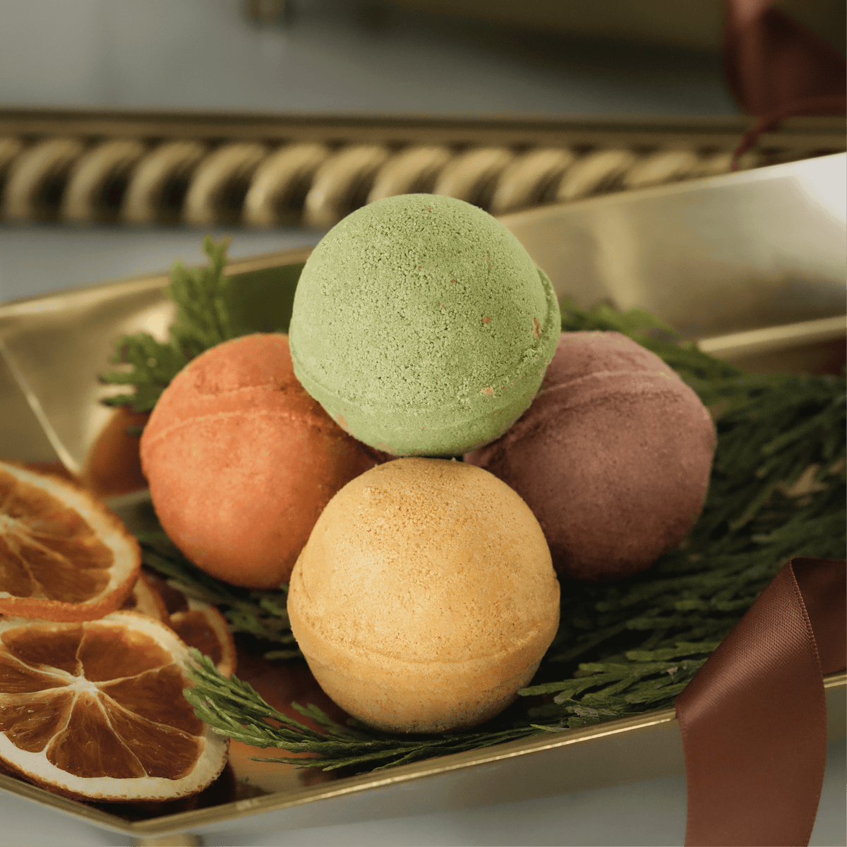 Four round bath bombs in green, peach, mauve, and tan, arranged on a gold tray with pine branches, dried orange slices, and a brown ribbon.