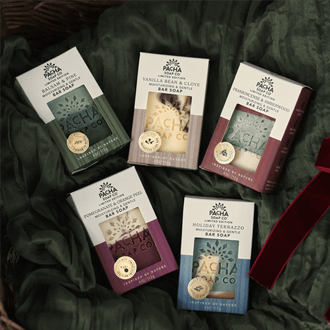 Five boxed bath soaps from Pacha Soap Co. arranged on dark green draped fabric, with a red ribbon at the lower right.