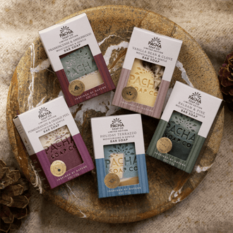 Five boxed Pacha Soap Co. handcrafted bar soaps arranged on a round wooden tray with a linen surface and pinecones.