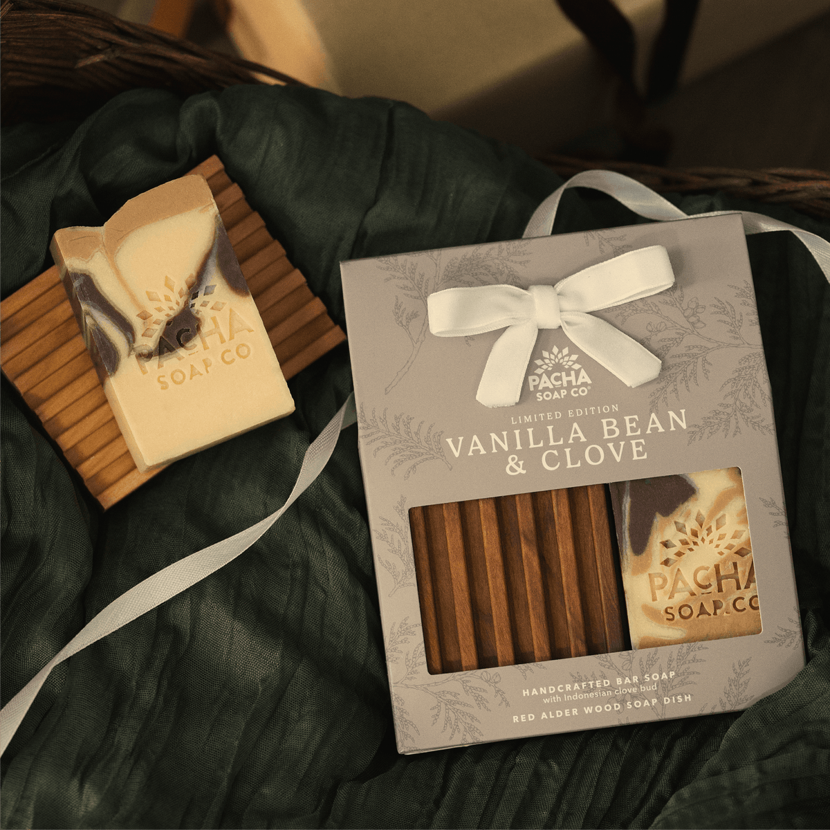 Pacha Soap's Creamy Beige Bar Soap & Dish Set package is sitting next to the barsoap and wooden dish that are in the set, sitting by ribbon and wrinkled green fabric. 