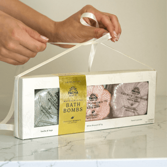 Two hands tying a white ribbon over Pacha Soap Co's Signature Bath bomb set with Vanilla & Sage, White Blossom & Fig, and French Lavender Bath bombs