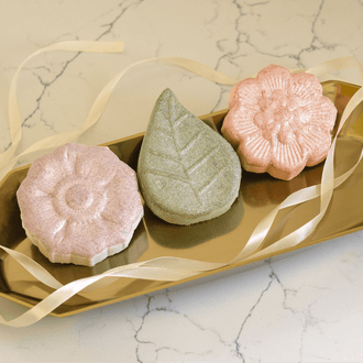 Pacha Soap Co's Signature Bath Bomb Set out of the box, with French Lavender, Vanilla & Sage, and White Blossom & fig lined up on a golden tray with white ribbon draped across it.