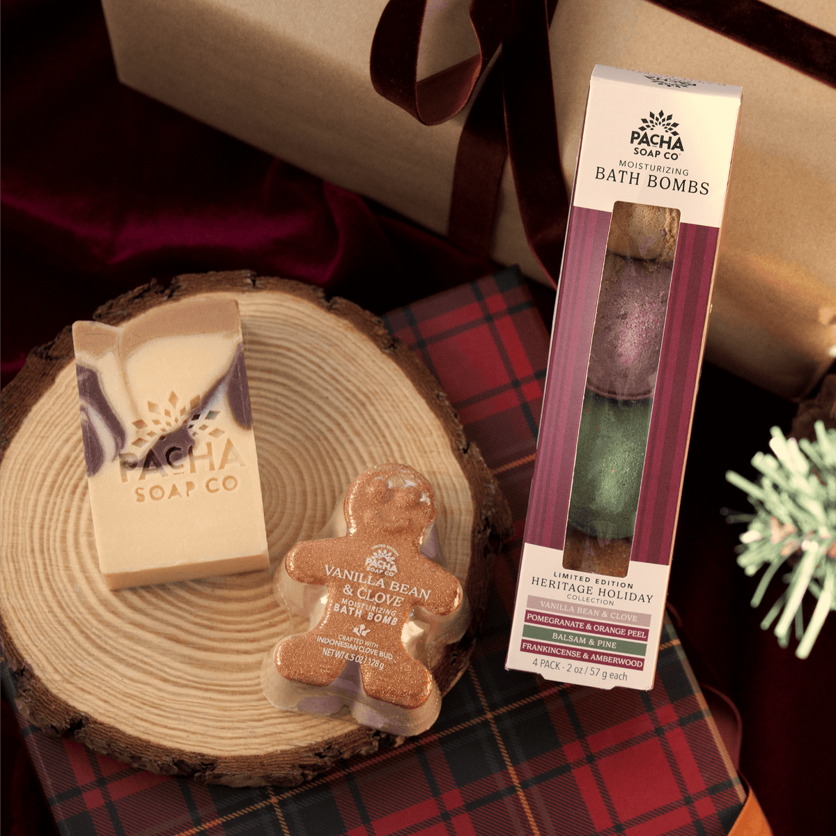 An assortment of Pacha Soap Co's Holiday Heritage items. A bar of Vanilla Bean & Clove soap, a set of the new scents as bath bombs, and a vanilla bean & clove bath bomb all gather on a wooden log tray and a plaid gift.