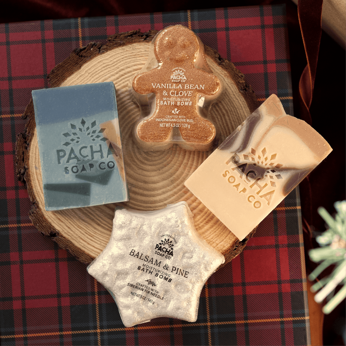 An assortment of Pacha Soap Co's Vanilla Bean & Clove's Bar Soap and Bath bomb, a holiday terrazzo bar soap and Balsam & Pine Bath Bomb on a wooden log tray and plaid gift