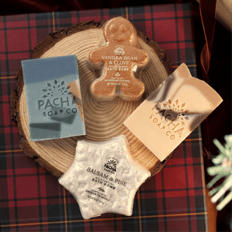 An assortment of Pacha Soap Co's Vanilla Bean & Clove's Bar Soap and Bath bomb, a holiday terrazzo bar soap and Balsam & Pine Bath Bomb on a wooden log tray and plaid gift