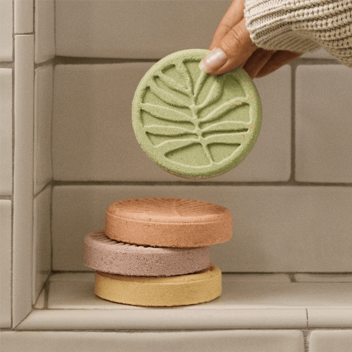 A hand wearing a cream knit sleeve holds a round light green shower steamer with a leaf embossed pattern above a stack of four round Shower Steamers in peach, mauve, and yellow, set against a beige tiled background.