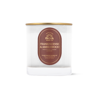 A clear glass jar candle with a gold metal lid and a red oval label reading 'PACHA SOAP CO FRANKINCENSE & AMBERWOOD'. 
